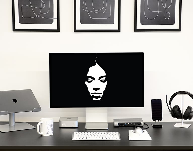Modern desk setup with Kensington keyboard, trackball, docking station, and headphones. A Mac Mini, MacBook on a stand, smartphone, and a monitor displaying a stylized face are also present. Abstract black-and-white art hangs above.