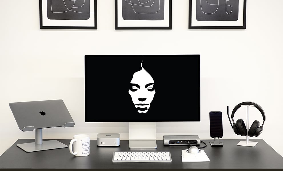 Modern desk setup with Kensington keyboard, trackball, docking station, and headphones. A Mac Mini, MacBook on a stand, smartphone, and a monitor displaying a stylized face are also present. Abstract black-and-white art hangs above.