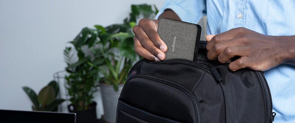 A person places a compact Kensington docking station into a black backpack.