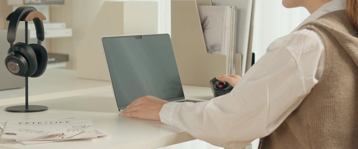 A person uses a laptop equipped with a Kensington privacy screen, holding a trackball mouse, in a well-organized workspace.