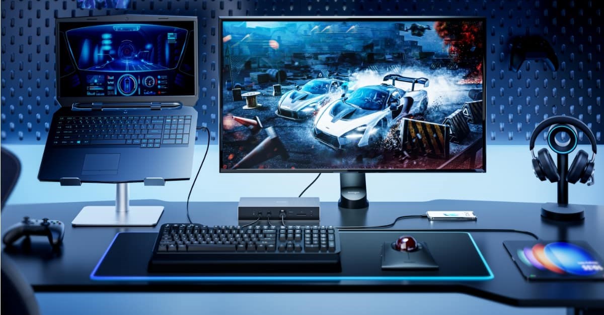 Gaming setup featuring a laptop on a stand, a large monitor displaying a car racing game, a keyboard, a trackball mouse and headphones on a desk.