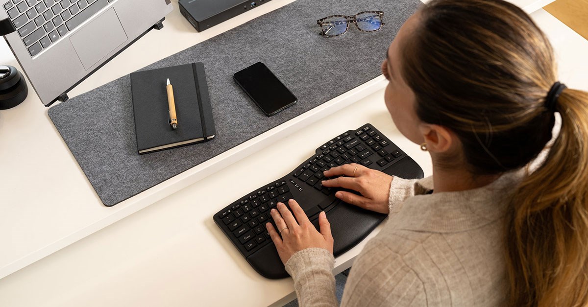 Woman sitting at a desk, typing on a Kensington ergonomic keyboard. The desk setup includes a laptop, notebook and smartphone.