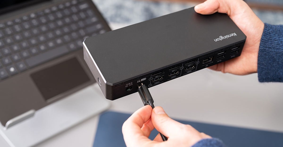 Hands connect a cable to a Kensington docking station with multiple HDMI and DisplayPort options near a laptop.