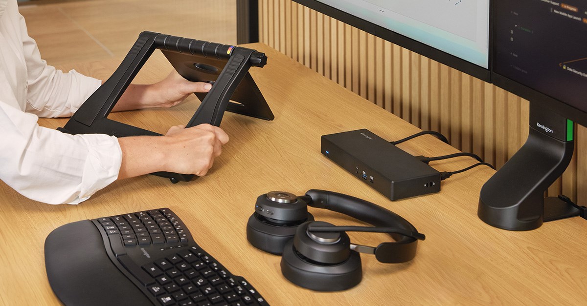 Hands adjust a black laptop riser on a desk, alongside a keyboard, headphones, and a docking station.