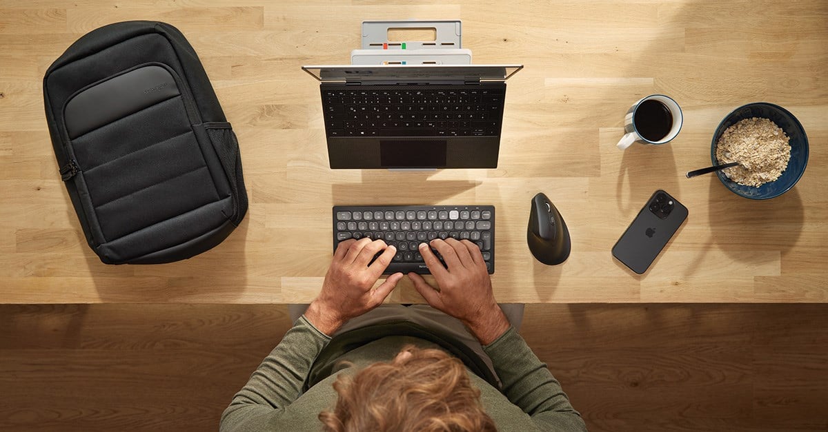 A person works at a wooden desk. The setup includes a Kensington laptop riser, a compact keyboard and an ergonomic vertical mouse.