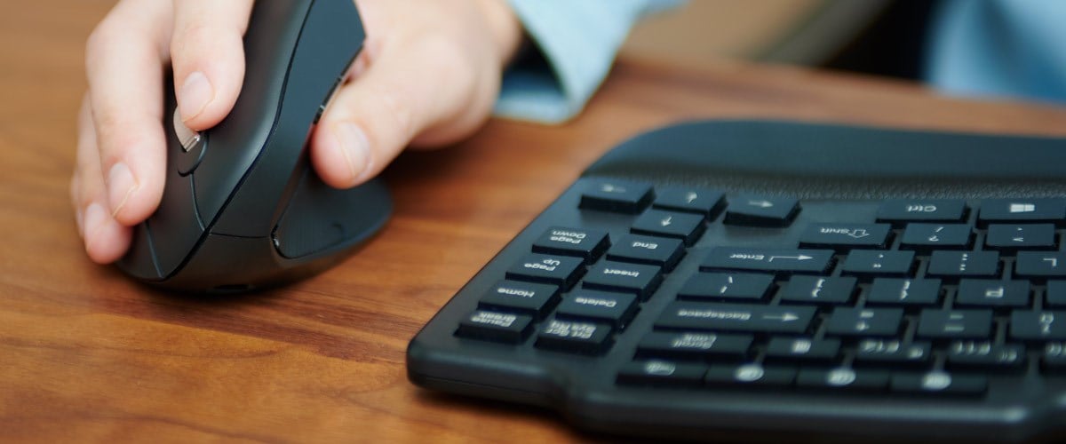 A close-up of a hand on a vertical ergonomic mouse next to a curved ergonomic keyboard.