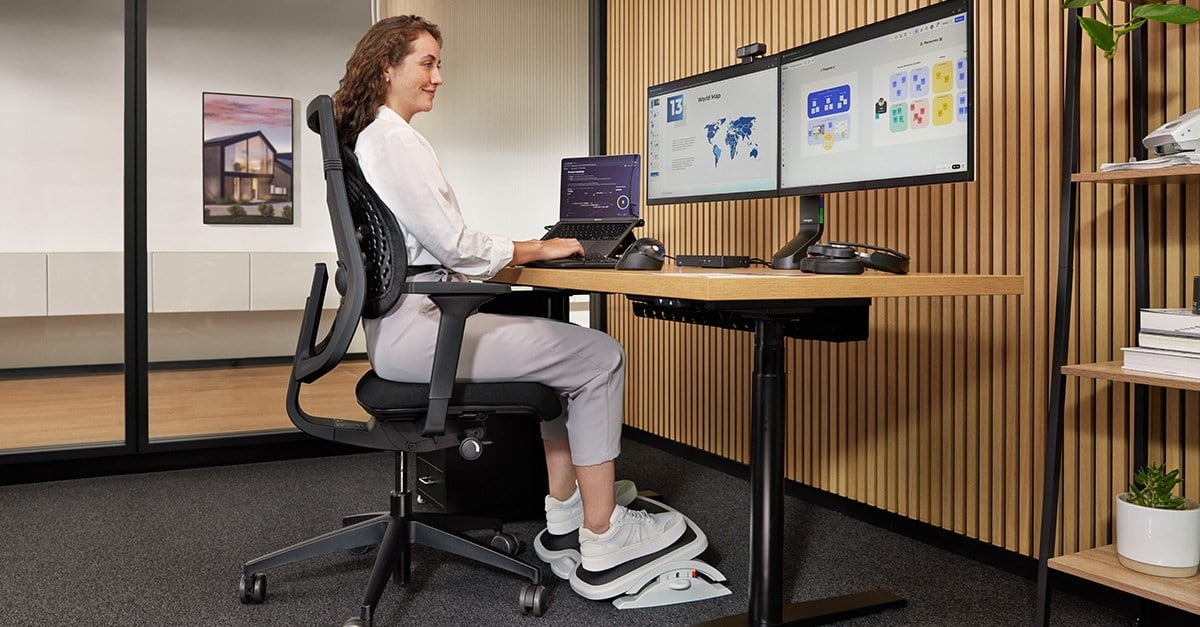 A woman sits at a desk using a laptop, ergonomic chair, and a Kensington footrest.