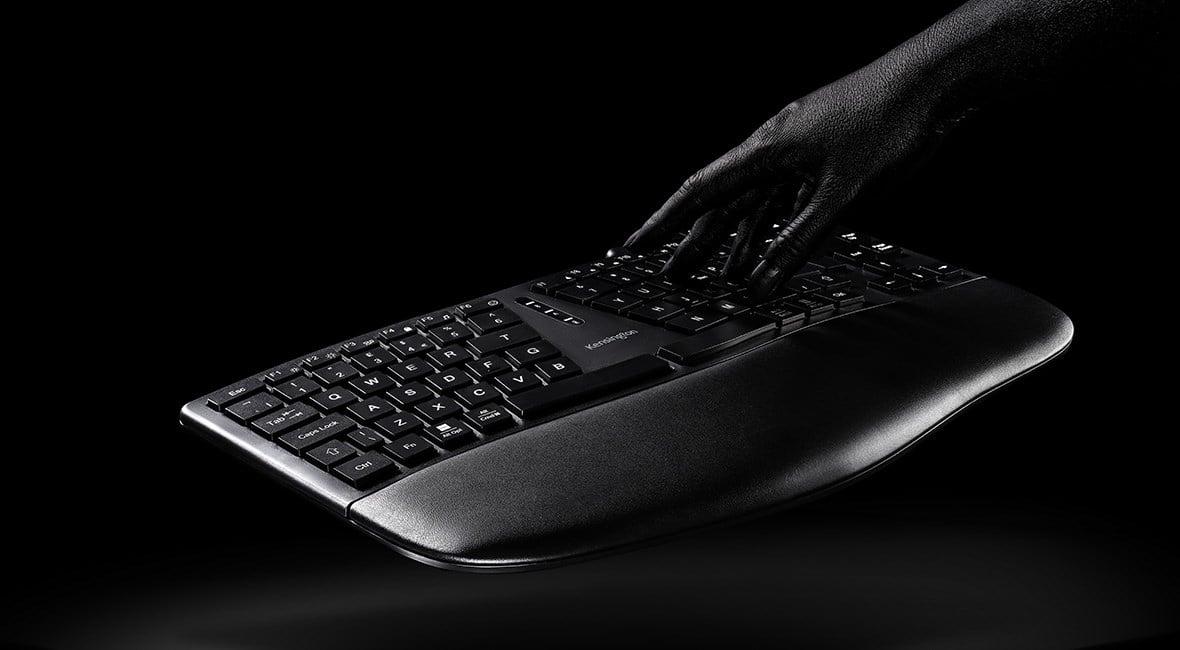 A black ergonomic Pro Fit Ergo KB675 EQ TKL Keyboard floating over a dark black surface with a human hand preparing to type.